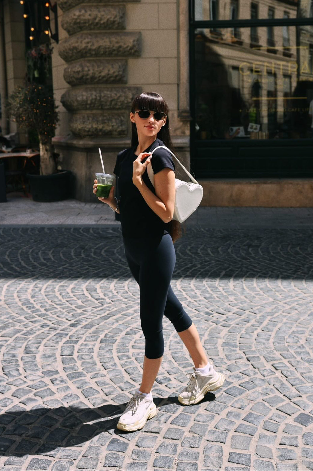 Woman walking on a cobblestone street holding a drink, wearing a black outfit and sunglasses.